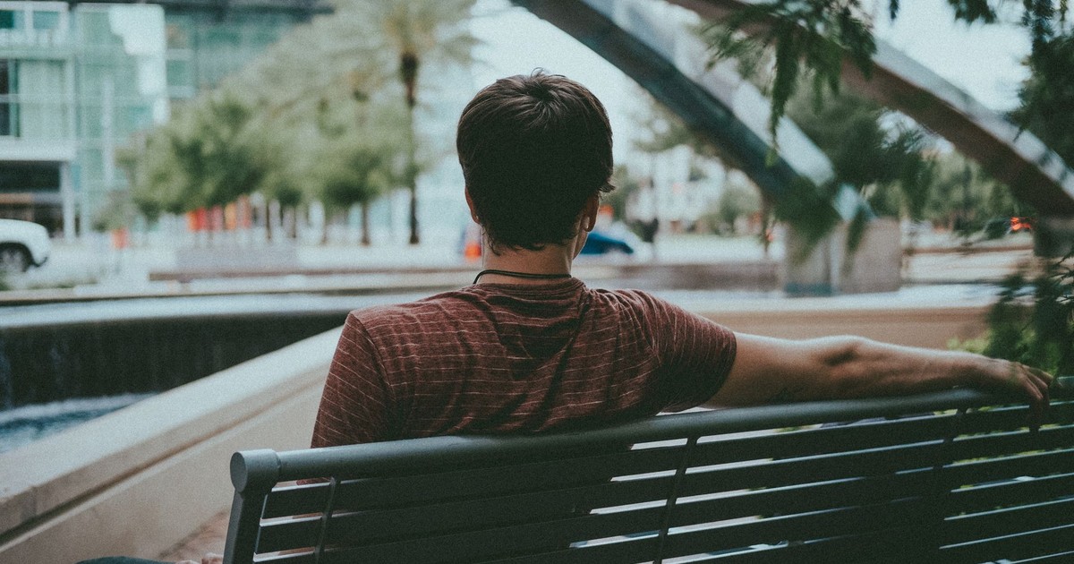 A man in a striped shirt sits and relaxes on a bench in an urban area, surrounded by greenery.