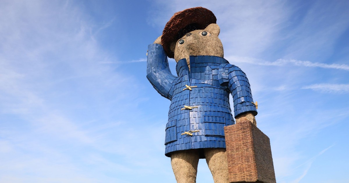 Large bear statue with blue coat and suitcase, outdoors under a clear sky in Hurleston, UK.