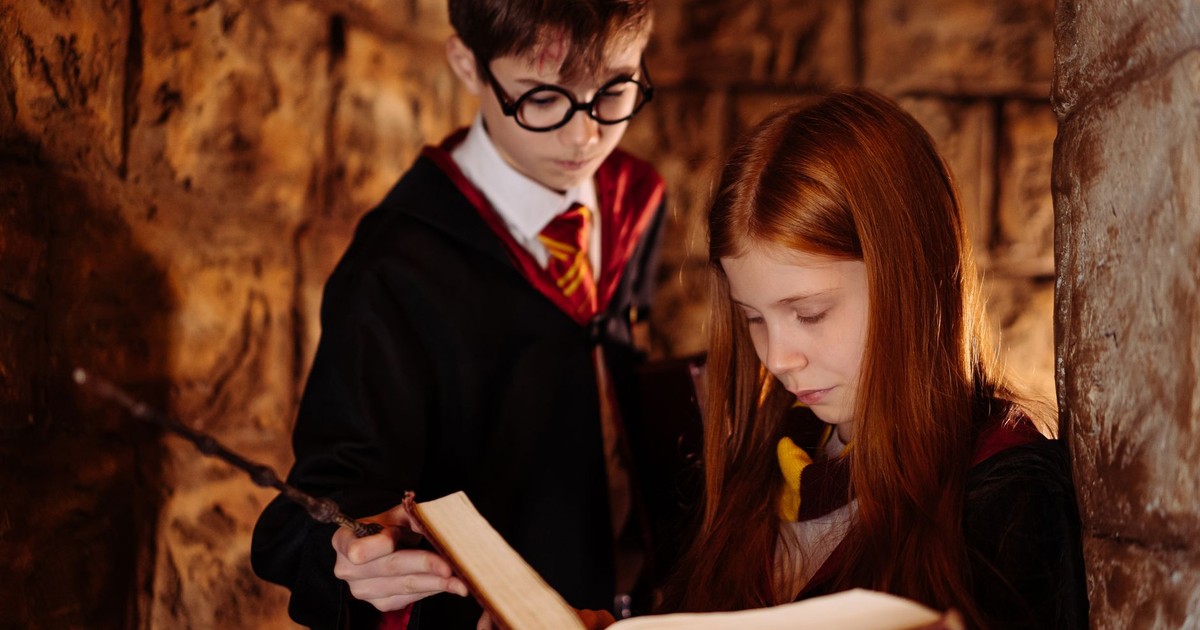 Two children in wizard costumes reading a spellbook in a dimly lit stone room.