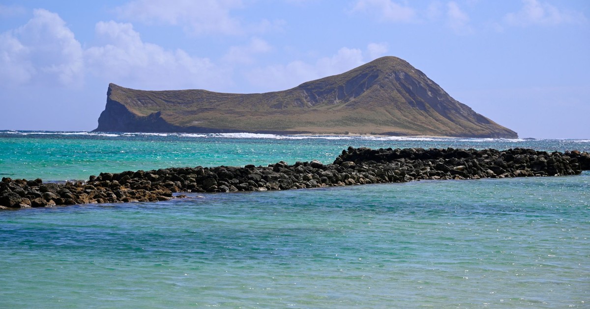 Scenic view of a distant island surrounded by aqua blue waters and stone barrier under a blue sky.