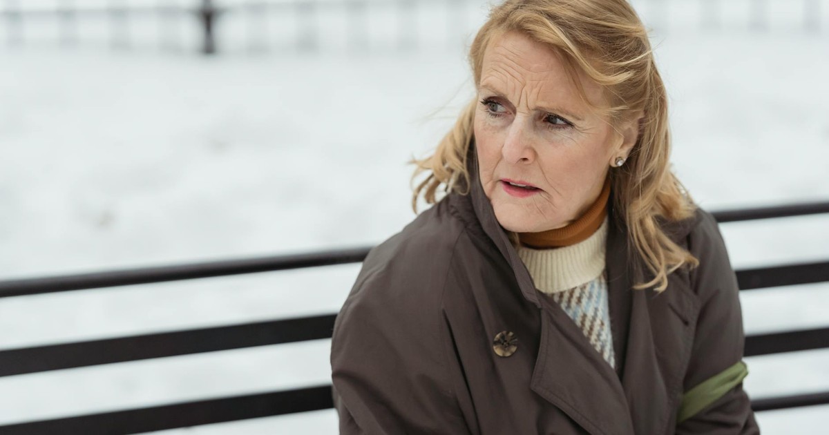 Contemplative elderly female with wrinkled skin in outerwear looking away while resting on urban bench in wintertime