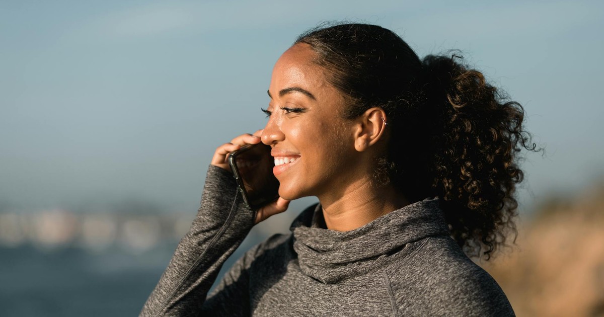 A smiling woman in a gray sweater enjoying a phone call outdoors by the sea.