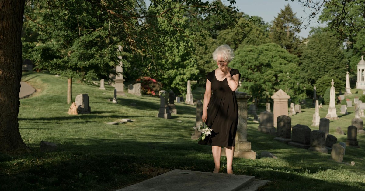 An elderly woman grieving by a grave with white flowers in a peaceful cemetery.