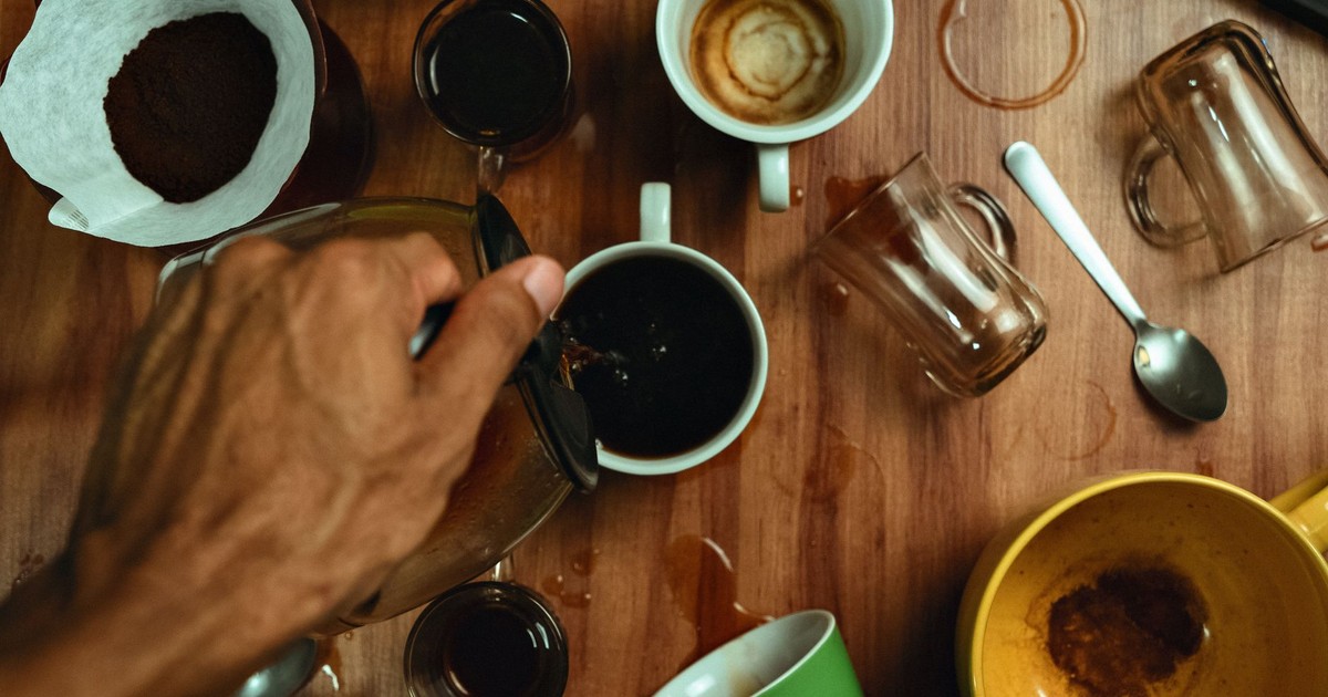 Top view of a hand pouring coffee into a mug surrounded by spilled cups on a wooden table.