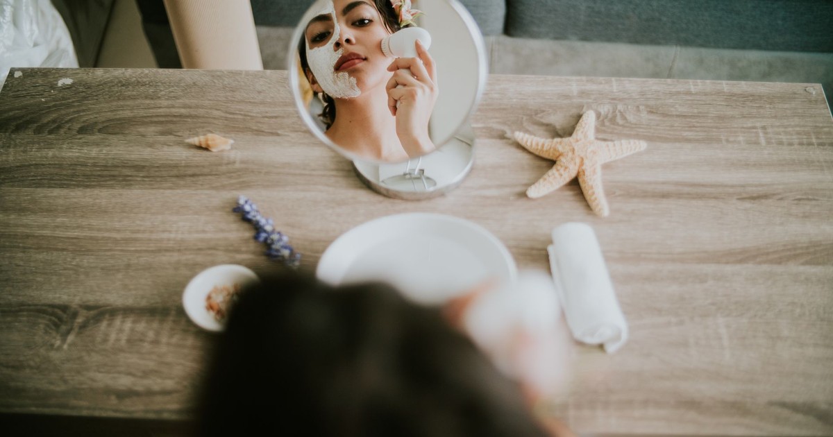 A woman applies a facial mask in front of a mirror with spa elements on a wooden table.
