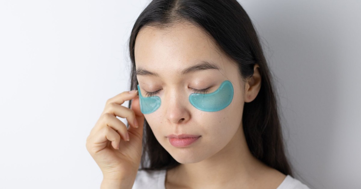 Serene Asian woman with closed eyes applying blue under-eye patches for skincare routine.