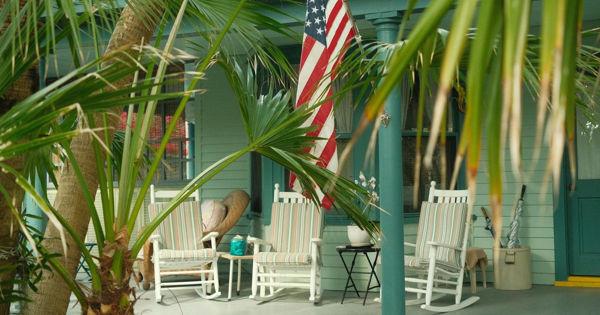 Charming Southern porch with American flag and rocking chairs, hidden behind lush palm trees.