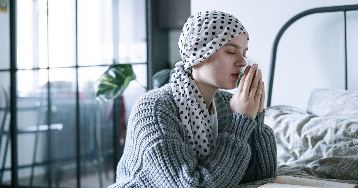 A hopeful woman in a scarf praying in a cozy bedroom during her cancer recovery journey.