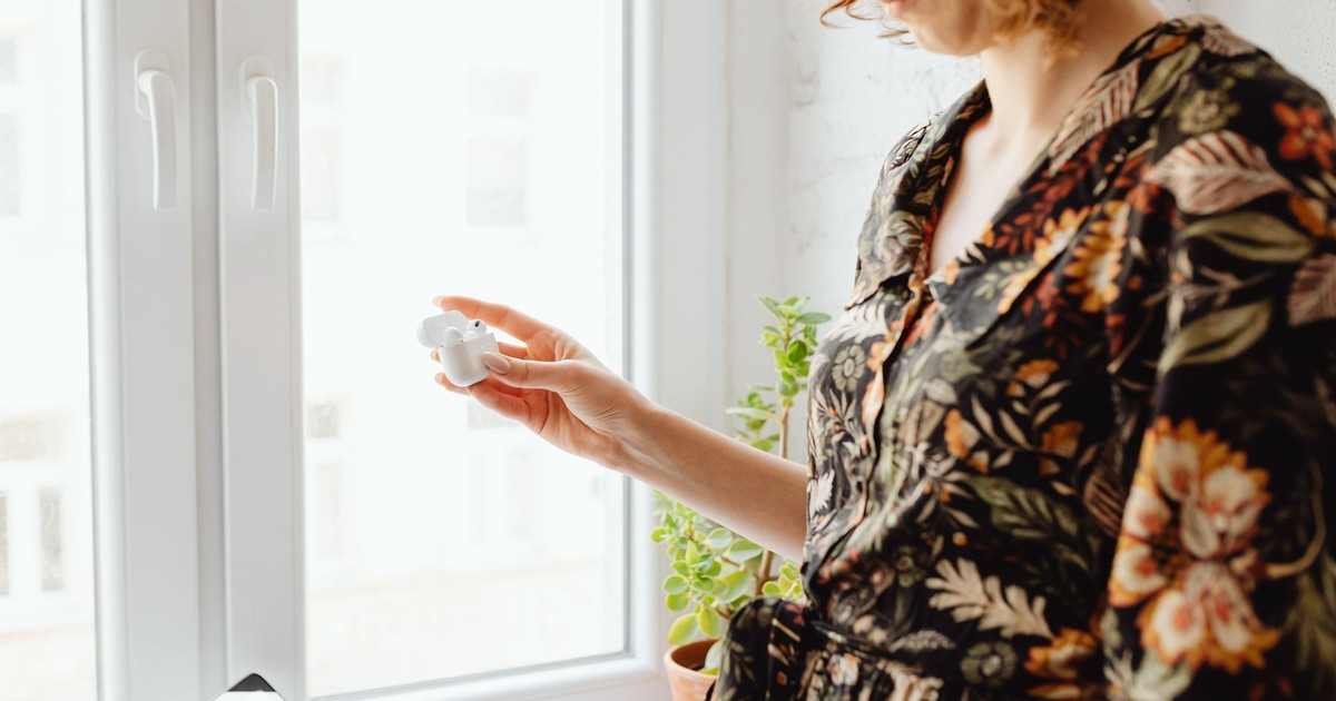 A woman stands by a window holding wireless earbuds, creating a modern lifestyle vibe.