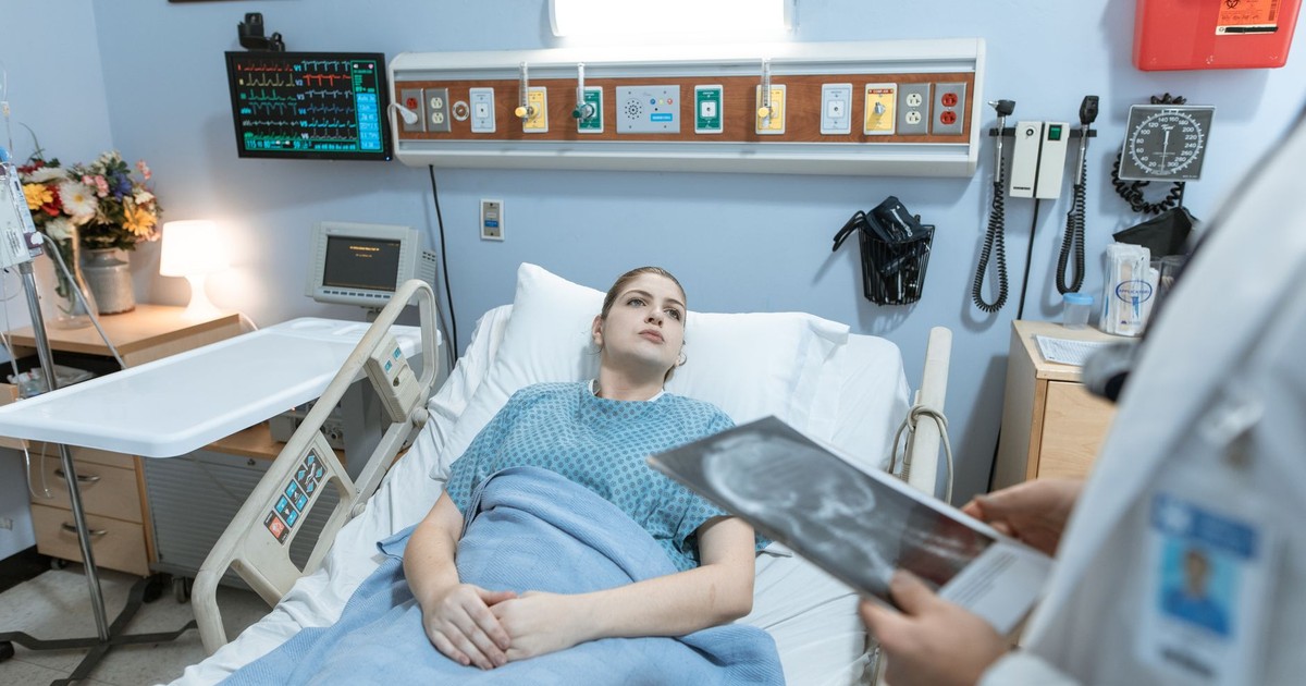 A female patient in a hospital gown receives medical attention from a healthcare professional holding an x-ray in a hospital ward.