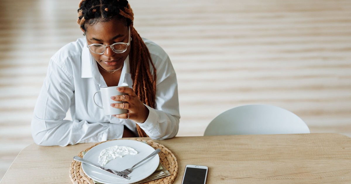 Woman in white shirt enjoying coffee at breakfast with smartphone in view.