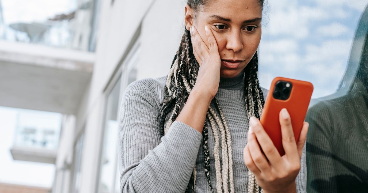 Crop serious African American female with dreadlocks touching cheek in contemplation and browsing mobile phone on street