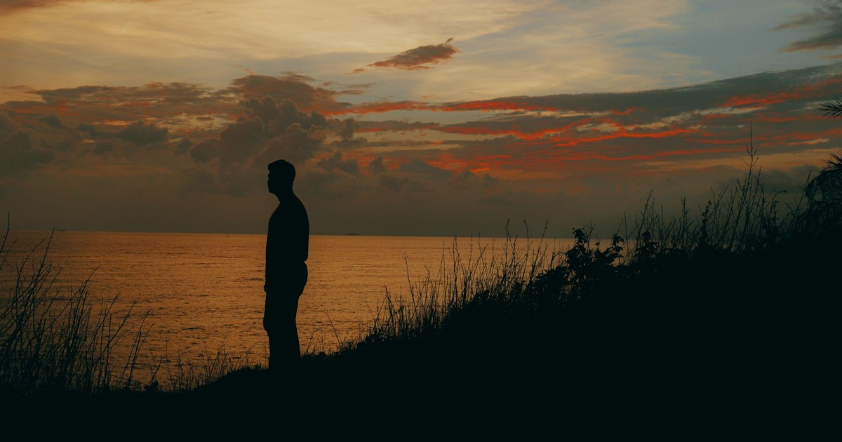 Silhouette of a person by the seaside during sunset with a dramatic sky, invoking tranquility and contemplation.