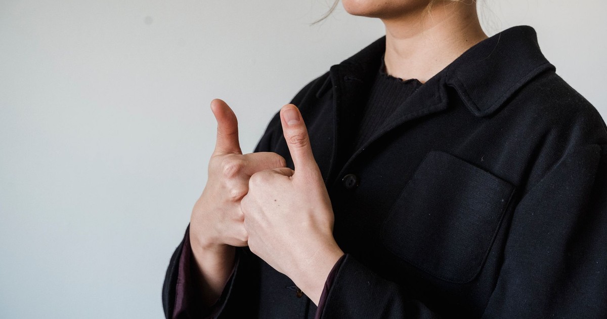 Close-up of a woman in a black jacket giving a double thumbs up gesture against a light background.