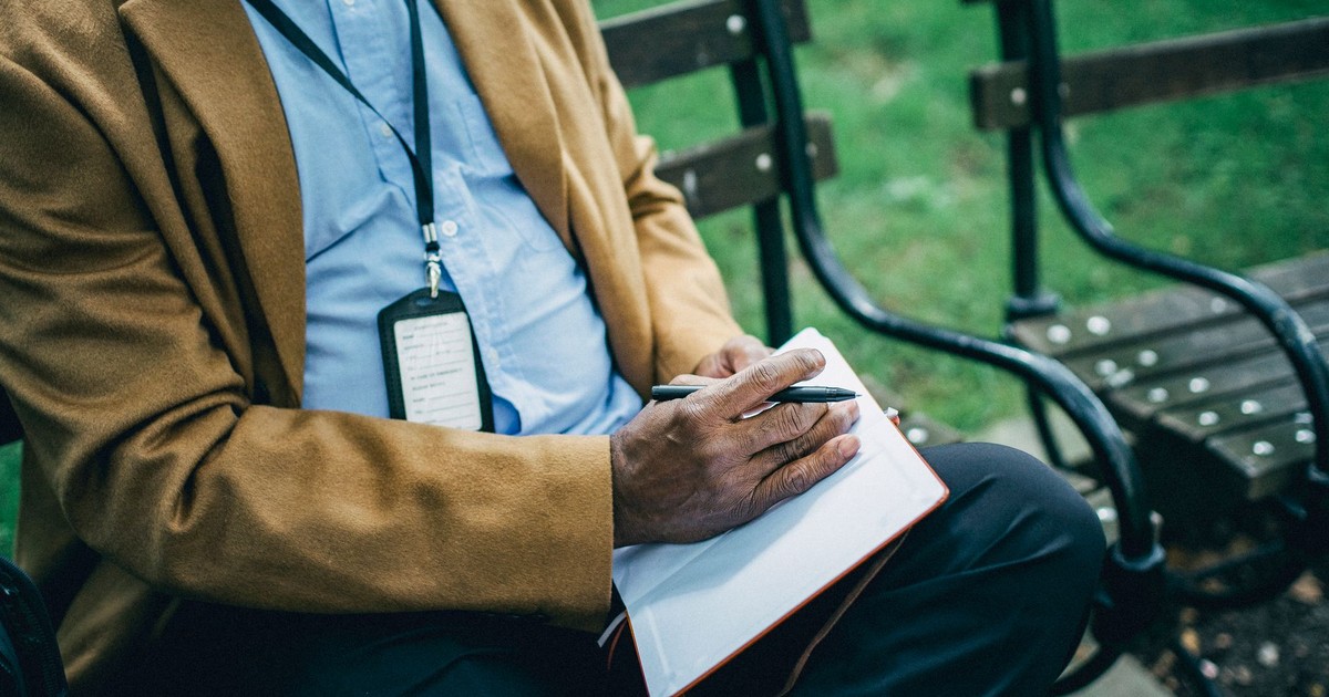 Crop anonymous African American male in formal wear sitting on wooden bench in park and taking notes in notebook
