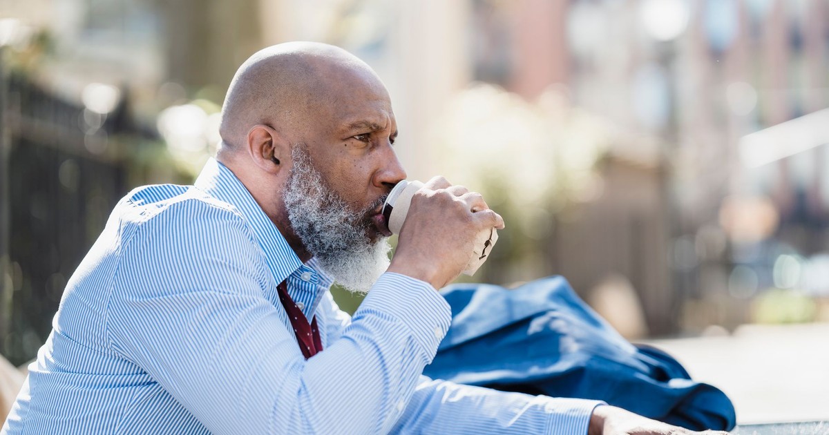 Senior man with beard relaxes while sipping coffee outdoors in a park setting.
