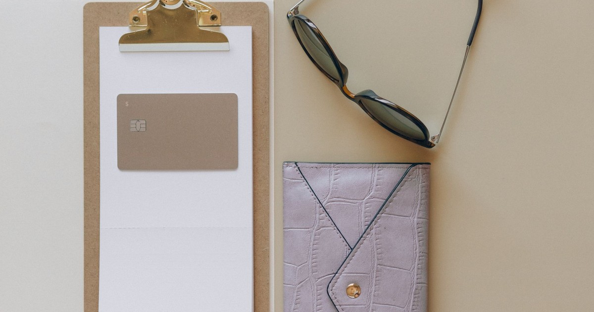 Flat lay image featuring a wallet, sunglasses, card, and clipboard on a beige surface.
