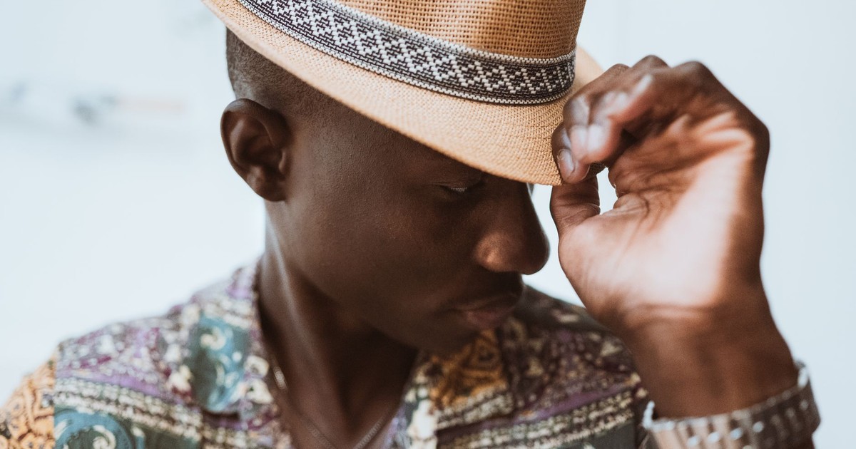 A fashionable man wearing a fedora and a colorful patterned shirt, adjusting his hat with a cool demeanor.
