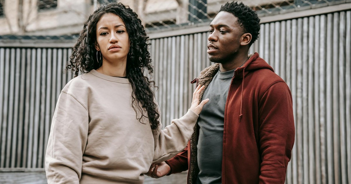 Sad ethnic girlfriend with curly hair rejecting annoyed African American boyfriend while arguing on street near wooden fence during breakup