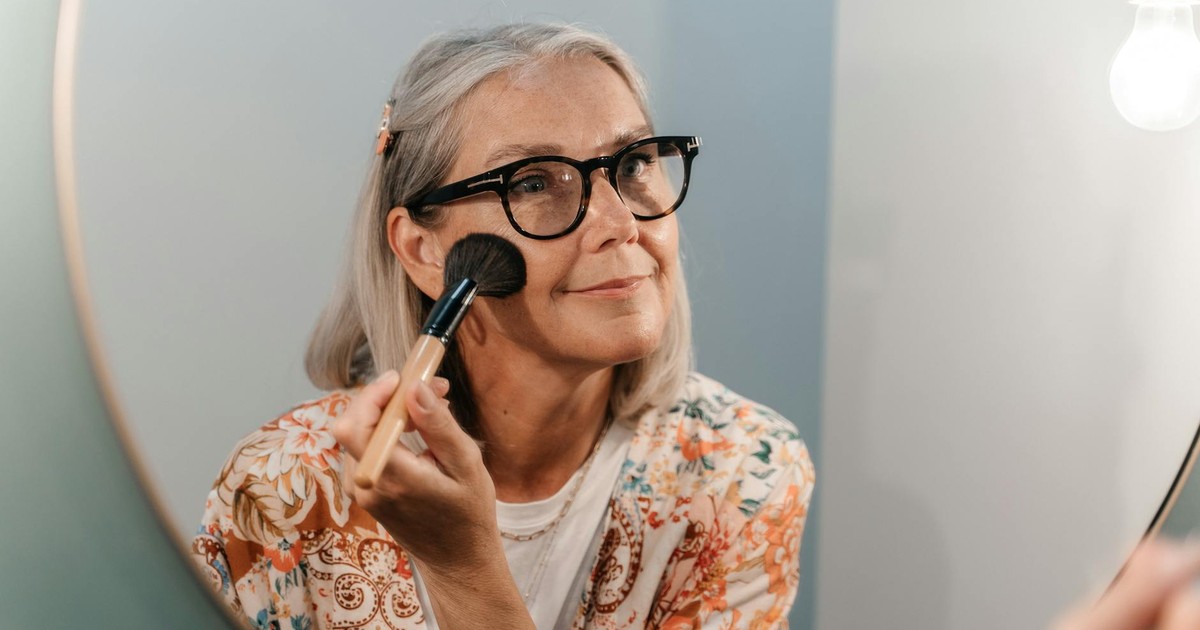 Senior woman with glasses applying makeup while looking in a mirror, reflecting a joyful moment.