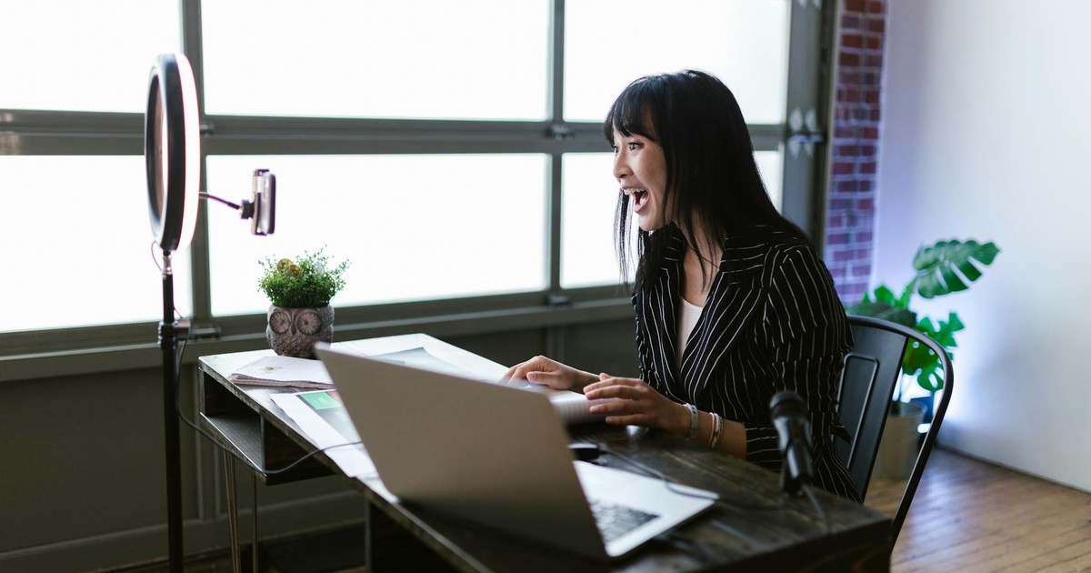Asian woman in office setting recording content with laptop and ring light, showcasing elegant business setup.