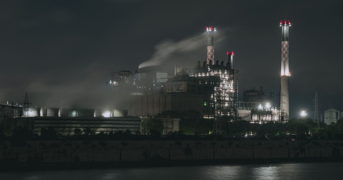 Night view of a large industrial factory with chimneys emitting smoke by the river in Ahmedabad, India.