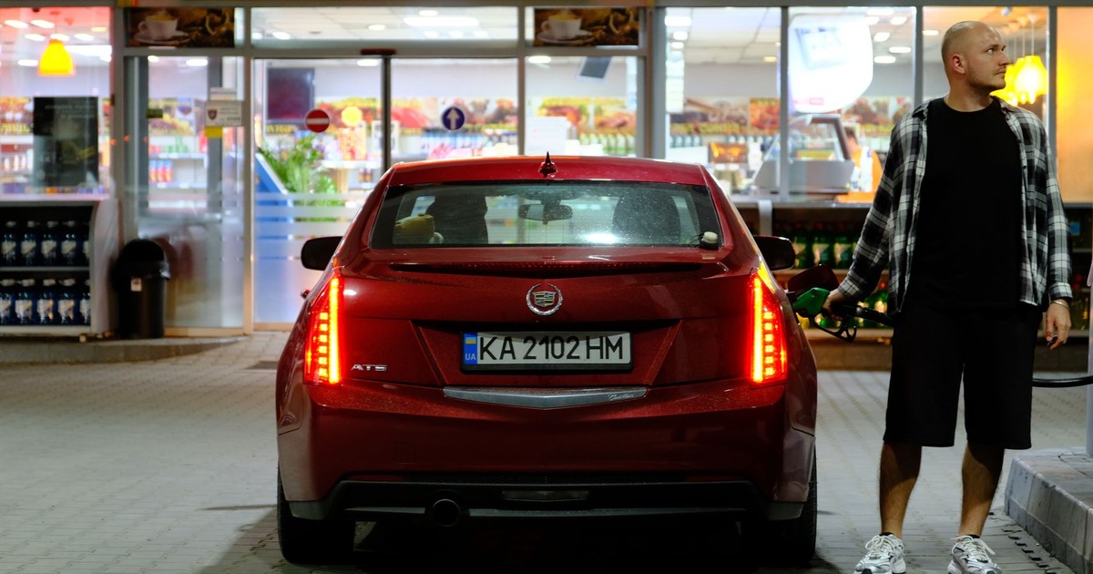 Man fueling a red car at a night-time gas station with a convenience shop in the background.
