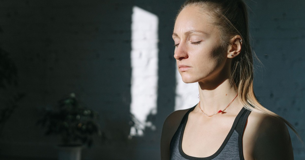 A woman in meditation inside a sunlit room, eyes closed, feeling calm and peaceful.