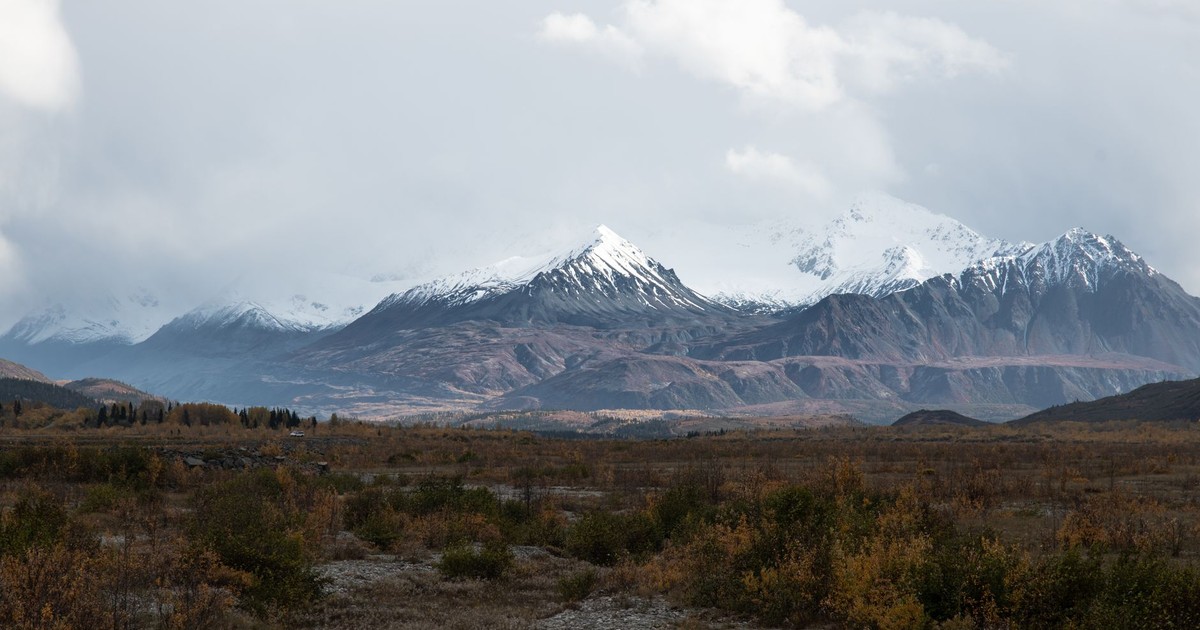 Breathtaking view of snow-covered Alaskan mountains with cloudy sky and autumn foliage in the foreground.