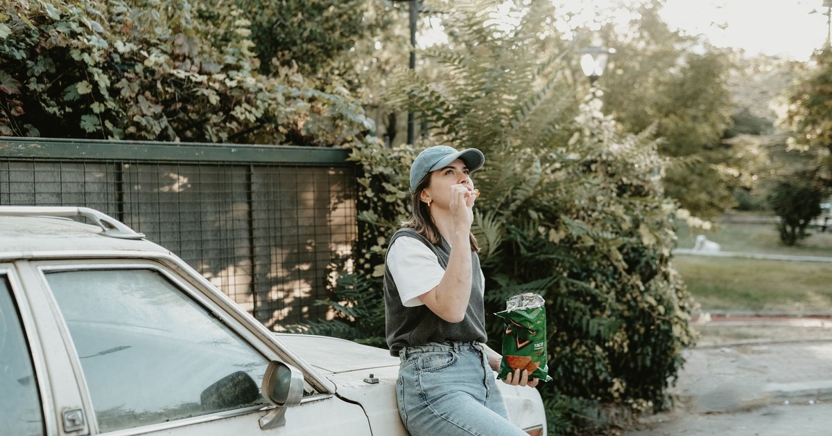 Woman in casual attire enjoys chips while leaning on a car outdoors on a sunny day.