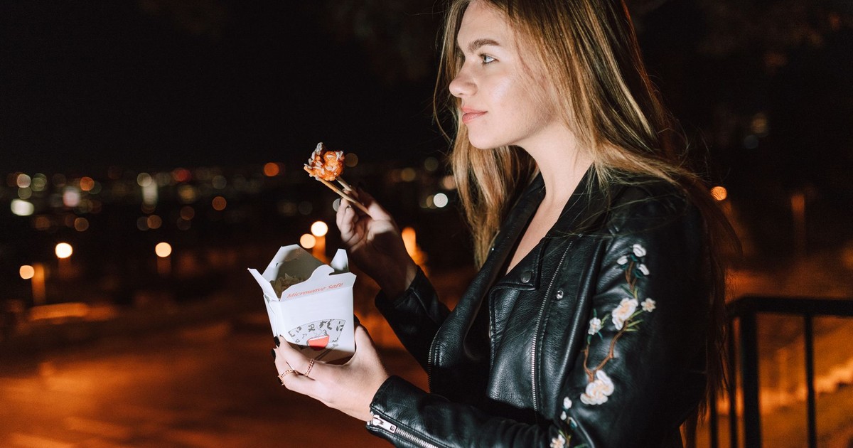 Young woman eating Asian takeout at night, city lights glowing in the background.