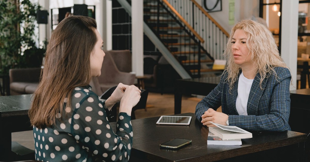 Businesswomen sitting at table with book and devices and having conversation about project in modern workplace