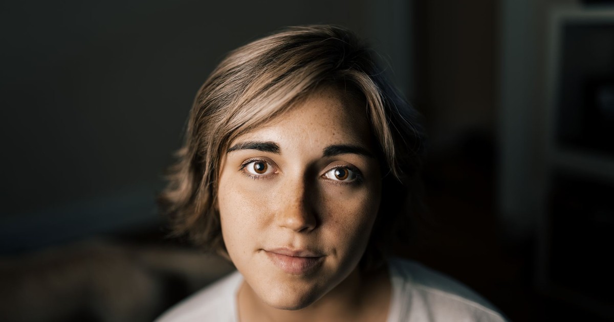 Young female with short hair in white t shirt looking at camera while standing in dark room with blurred background inside