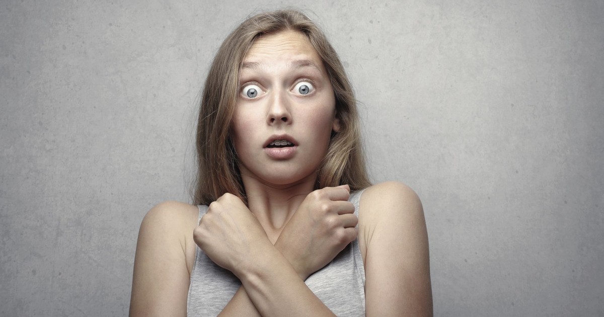 Portrait of a surprised woman with wide eyes and crossed arms against a gray background.