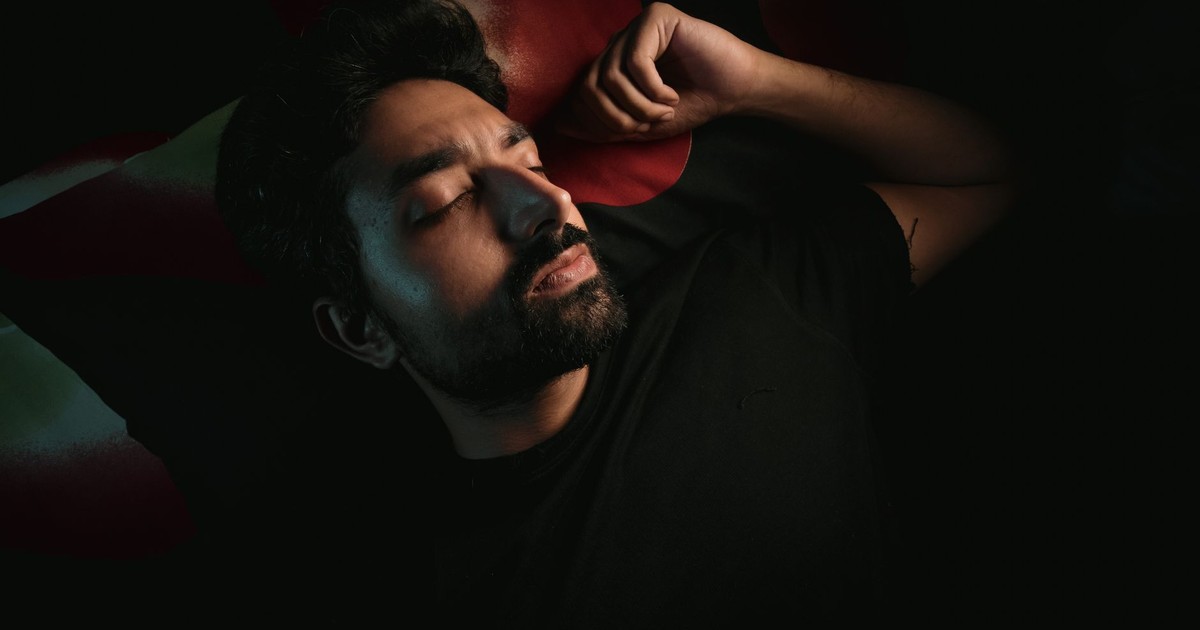 A bearded man lies in a dimly lit room, peacefully resting with closed eyes.