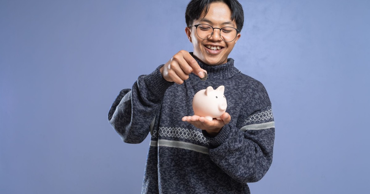 Smiling man holding a piggy bank and inserting a coin, representing savings and financial planning.