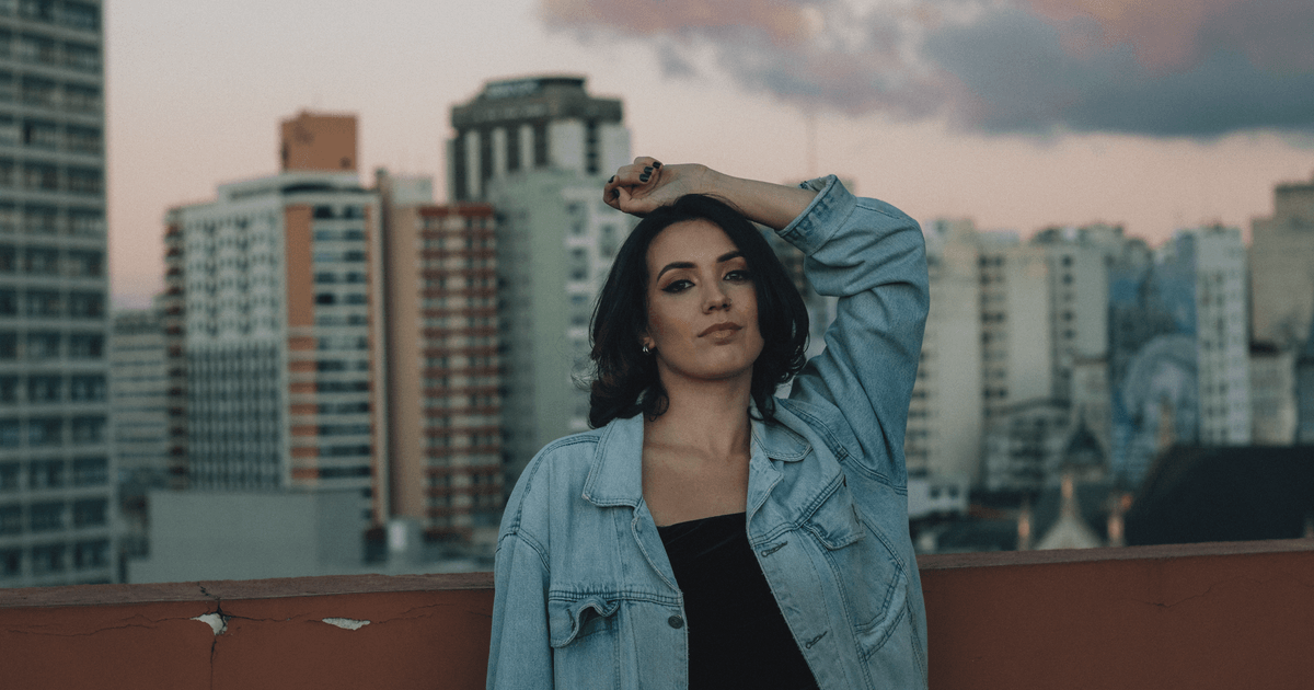 Stylish woman in denim jacket poses on a rooftop, cityscape at twilight.
