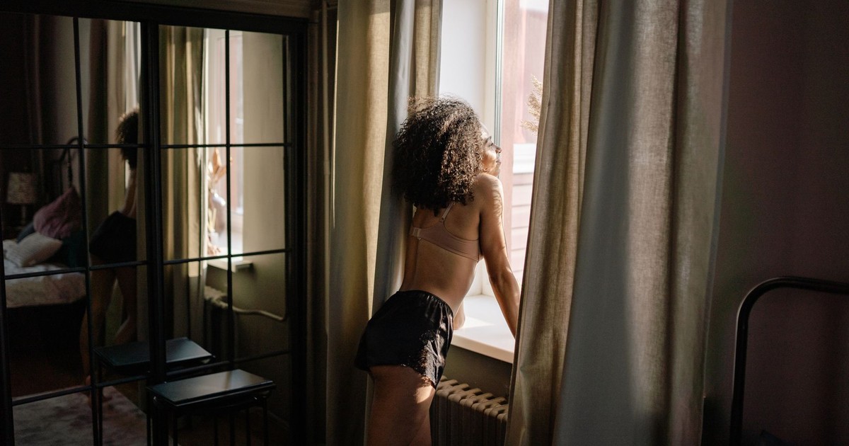 A woman in lingerie gazes through a window in a stylish living room setting.