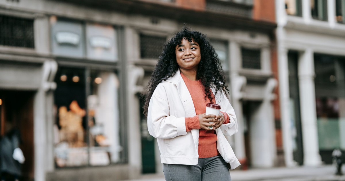 Smiling adult African American female in casual clothes with coffee to go walking on city street and looking away contentedly
