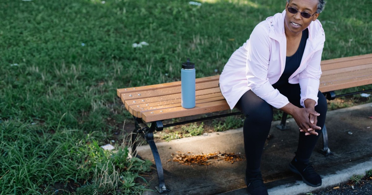Elderly woman sitting on a park bench with a tumbler, enjoying a peaceful day outdoors.