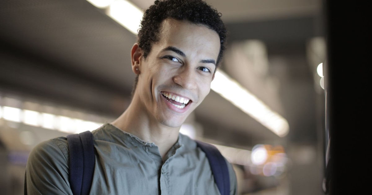 Low angle of joyful charismatic African American male in gray shirt with backpack smiling at camera while standing against blurred interior of contemporary building