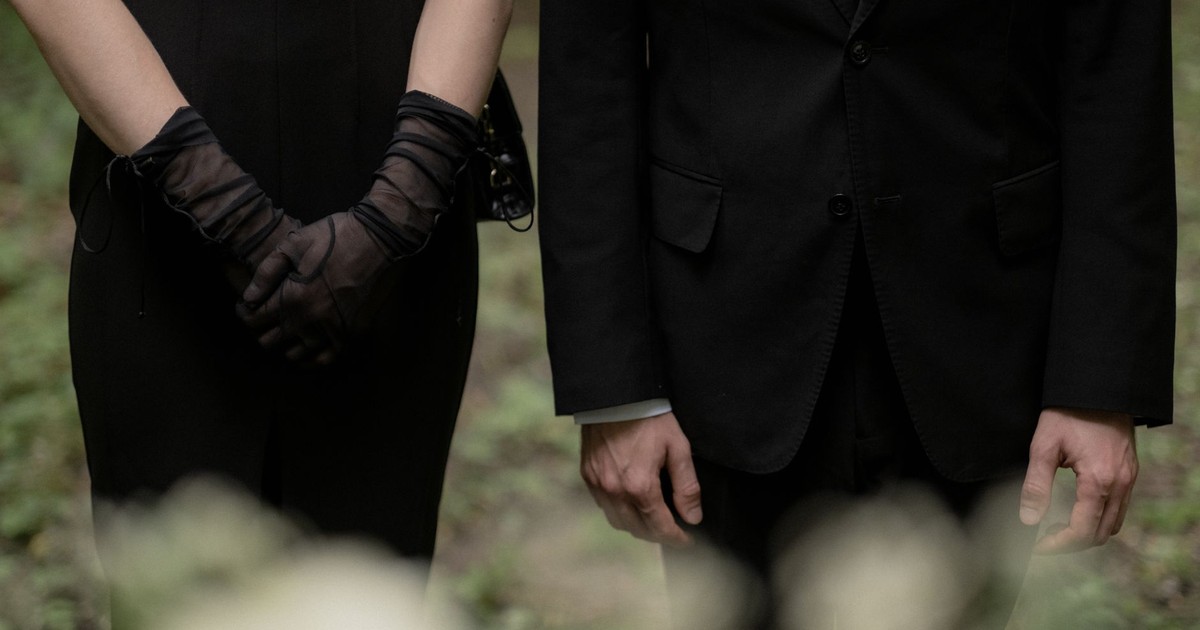 A man and woman in black formal wear stand side by side, exuding a somber mood.