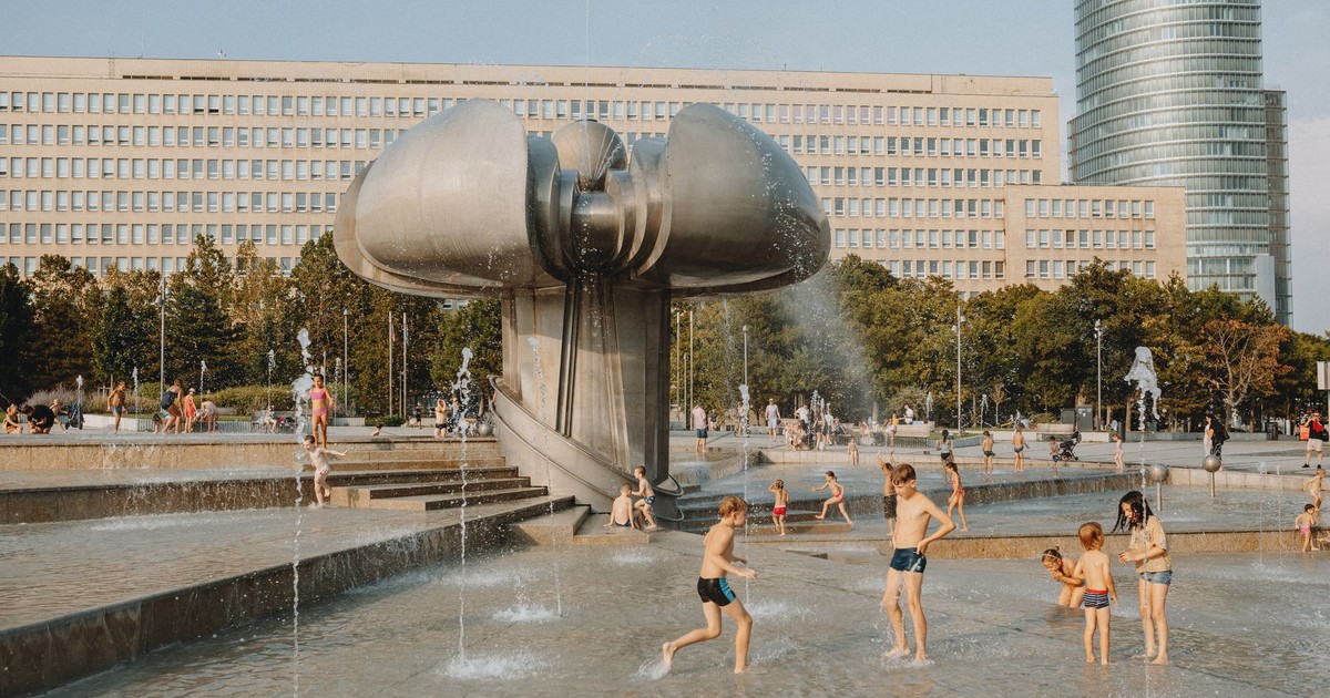 Kids enjoying a fountain in a city square with a sculpture and skyscraper in the background.