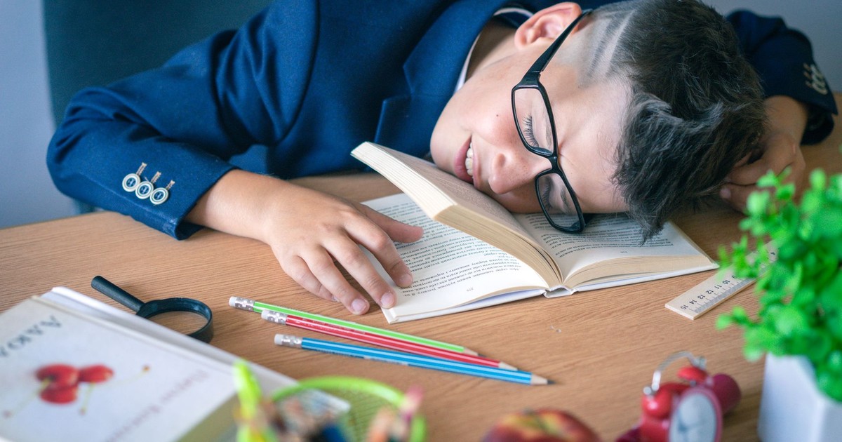 A tired schoolboy in glasses sleeping on a desk surrounded by study materials.
