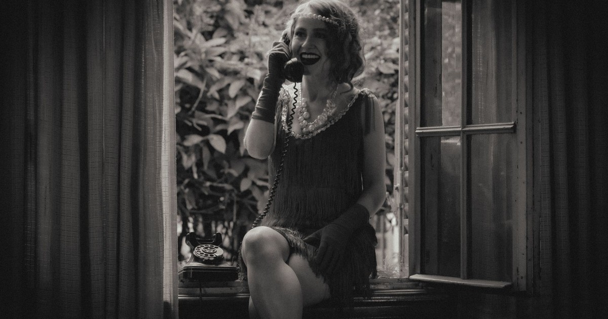 A woman in flapper attire sits by a window using a vintage telephone.