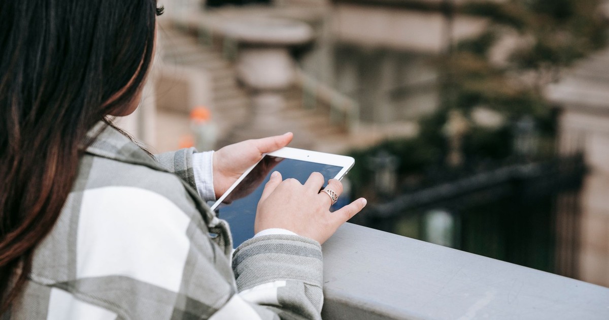From above of crop unrecognizable young female in checkered shirt text messaging on tablet while standing near railing