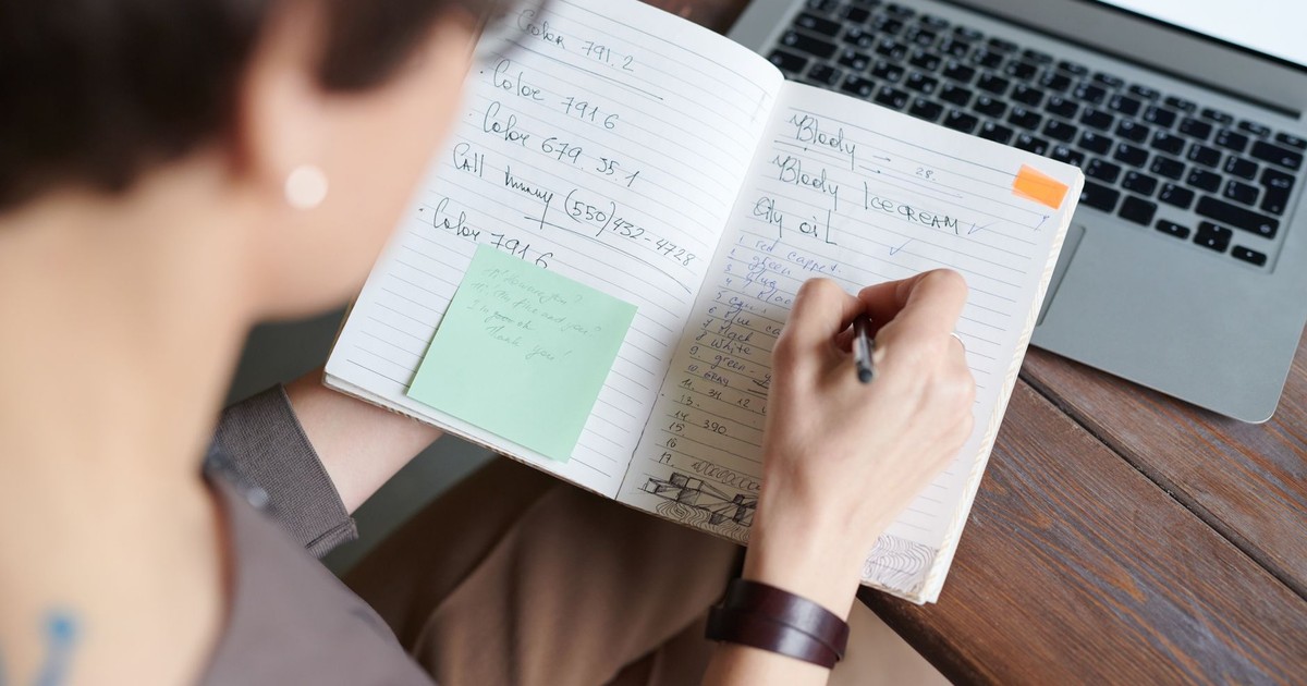 An adult writing in a notebook with a laptop on a wooden table for work or leisure.