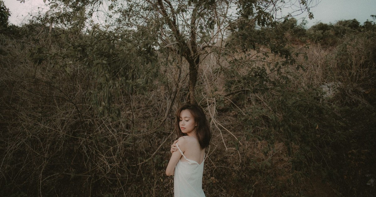 A young woman in a white dress stands peacefully in a natural, wooded area.