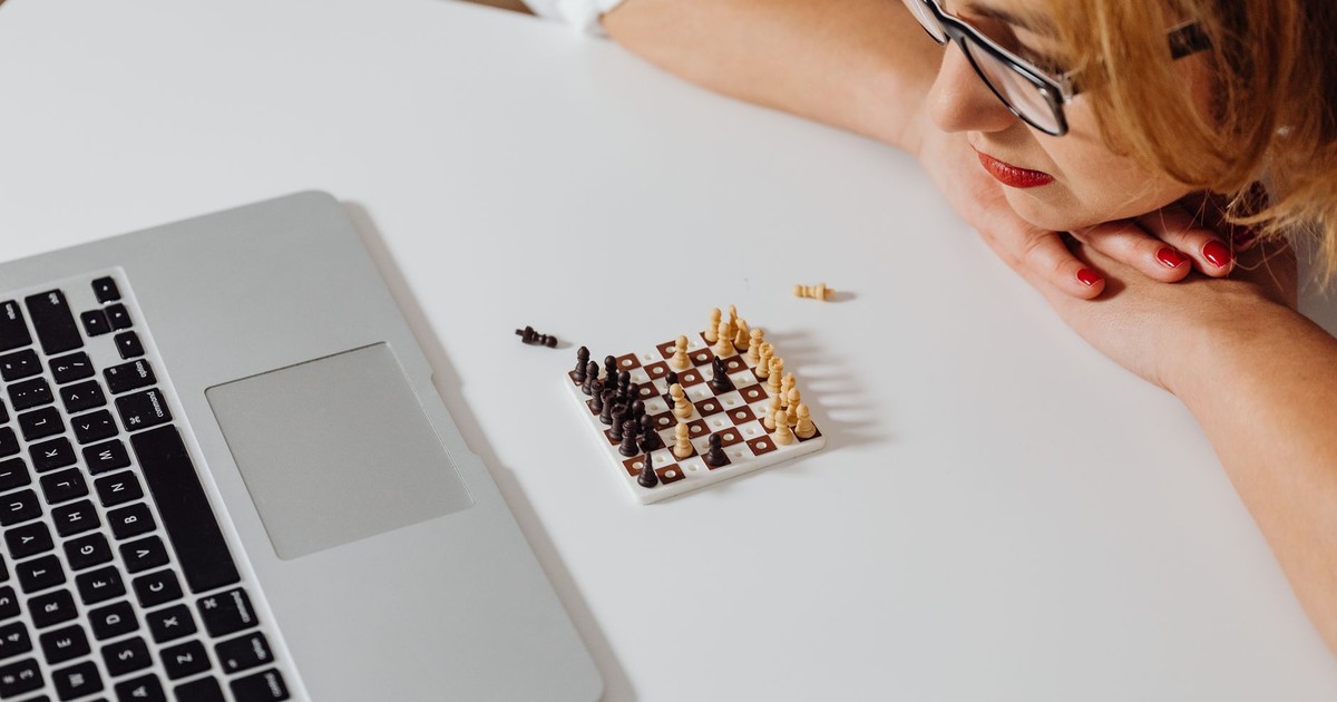 Focused woman looking at miniature chess set on white table with laptop nearby.