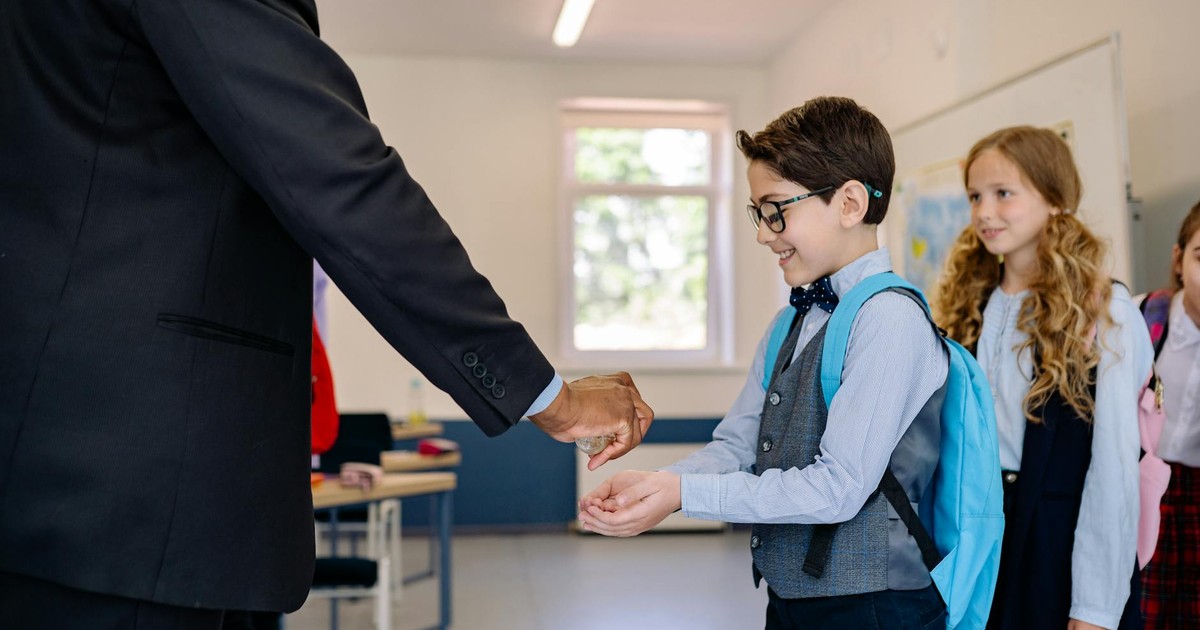 Children receiving hand sanitizer from a teacher in a classroom setting.